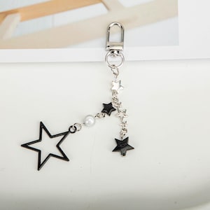 Może przedstawiać: Srebrny brelok do kluczy z motywem gwiazdy. Posiada dużą, ażurową czarną gwiazdę, perłę i grupę mniejszych gwiazd w kolorze czarnym i srebrnym. Brelok posiada zapięcie ułatwiające mocowanie.