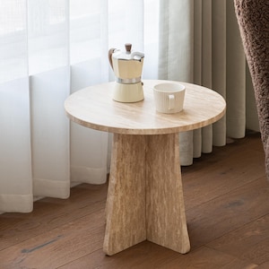 May include: A round beige travertine side table with a thick pedestal base. On the table surface sits a cream-colored moka pot with a brown handle and a white ribbed ceramic mug. The table features a natural porous stone texture throughout.