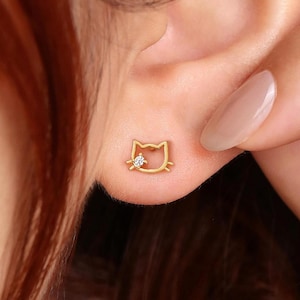 Może przedstawiać: Złote kolczyki sztyfty w kształcie kota, każdy z małym, przezroczystym kamieniem szlachetnym. Kolczyki są noszone na uchu, a dłoń delikatnie dotyka ucha. Tło jest w kolorze miękkiego różu.