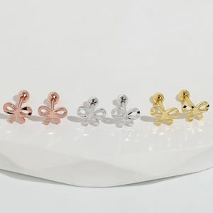 以下が含まれることがあります： ローズゴールド、シルバー、ゴールドトーンの、リボン型のスタッドピアスコレクション。各イヤリングは上部に小さなボールと繊細なリボンデザインが特徴です。イヤリングは白い表面に表示されています。