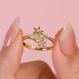 May include: A gold-colored ring featuring a star and a shooting star design. The ring has a delicate band and is adorned with small, clear gemstones. The ring is held against a pink background.