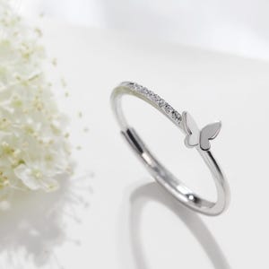May include: A silver ring with a butterfly design. The ring features a band with small, clear stones and a butterfly accent. The ring is displayed on a white surface with white flowers in the background.