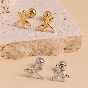 May include: Two pairs of dragonfly stud earrings with ball backings. One pair is gold-toned and the other is silver-toned. Each earring features a detailed dragonfly design with wings spread. The logo CRAZYCUTE THINGS is visible in the corner.