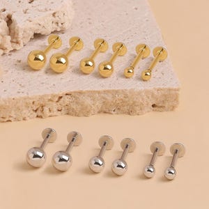 May include: A collection of twelve barbell earrings featuring smooth, polished spherical ends. Six of the earrings are finished in a bright gold tone, while the other six are finished in a polished silver tone, arranged in pairs of varying sizes.
