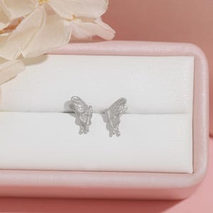 May include: Silver butterfly stud earrings presented in a pink jewellery box. The earrings feature intricate wing detailing. The background is a soft pink, with white flowers in the upper left corner.