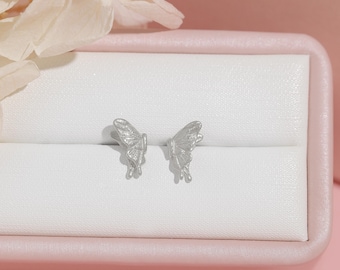 Könnte beinhalten: Ein Paar silberne Schmetterlings-Ohrstecker, präsentiert in einer rosa Schmuckschatulle. Die Ohrringe weisen filigrane Flügeldetails auf. Der Hintergrund ist ein zartes Rosa, mit weißen Blumen in der oberen linken Ecke.