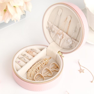 May include: A pink, round jewelry organizer with a zipper, open to reveal rings, necklaces, and hair accessories. The interior is lined with a soft, cream-colored fabric. Gold-colored jewelry and hair clips are visible, along with silver necklaces. The organizer is on a white surface.