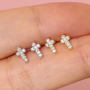 May include: Four small cross-shaped stud earrings. Two are silver-colored, and two are gold-colored. Each cross is encrusted with small, clear gemstones. The earrings are displayed against a light pink background.