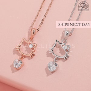 Könnte beinhalten: Zwei zarte Halsketten mit einem niedlichen Katzenanhänger und einem baumelnden Herzanhänger. Der Katzenanhänger hat einen kleinen Kristall in der Mitte und der Herzanhänger einen größeren Kristall. Eine Halskette ist silber und die andere ist roségold.