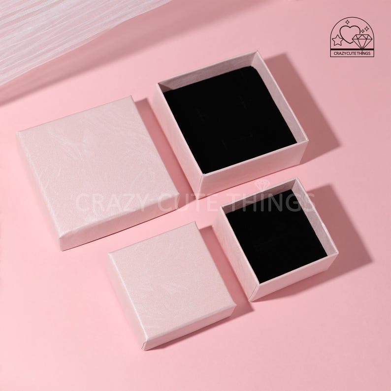 May include: Four pink square boxes with black velvet interiors. The boxes are of different sizes and are arranged in a staggered pattern on a pink surface. The boxes are ideal for storing small items such as jewellery or other trinkets.