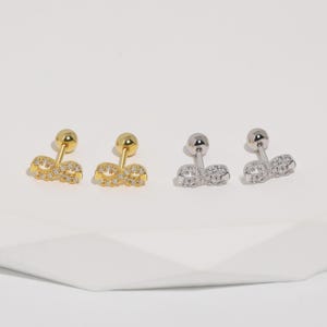 Op de afbeelding: Twee paar oorstekers. Eén paar is goudkleurig, het andere zilverkleurig. Elke oorbel heeft een kleine, ronde stud en een decoratief, dubbel strik-ontwerp bezet met kleine, heldere kristallen. De oorbellen worden gepresenteerd op een wit oppervlak.