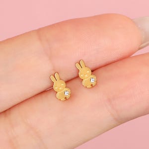 May include: A pair of gold-colored bunny stud earrings. Each earring features a small, stylized bunny with a sparkling crystal accent on its belly. The earrings are set against a soft pink background.