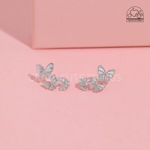 May include: A pair of silver butterfly earrings with clear crystal accents. Each earring features three butterflies, with the largest butterfly at the back and two smaller butterflies in front.