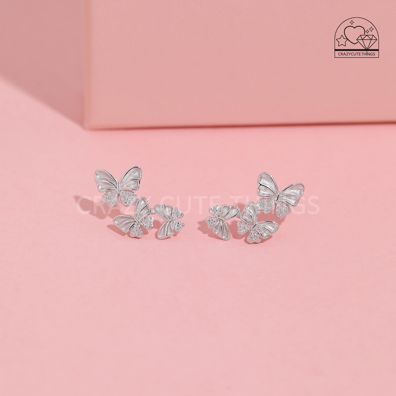 May include: A pair of silver butterfly earrings with clear crystal accents. Each earring features three butterflies, with the largest butterfly at the back and two smaller butterflies in front.