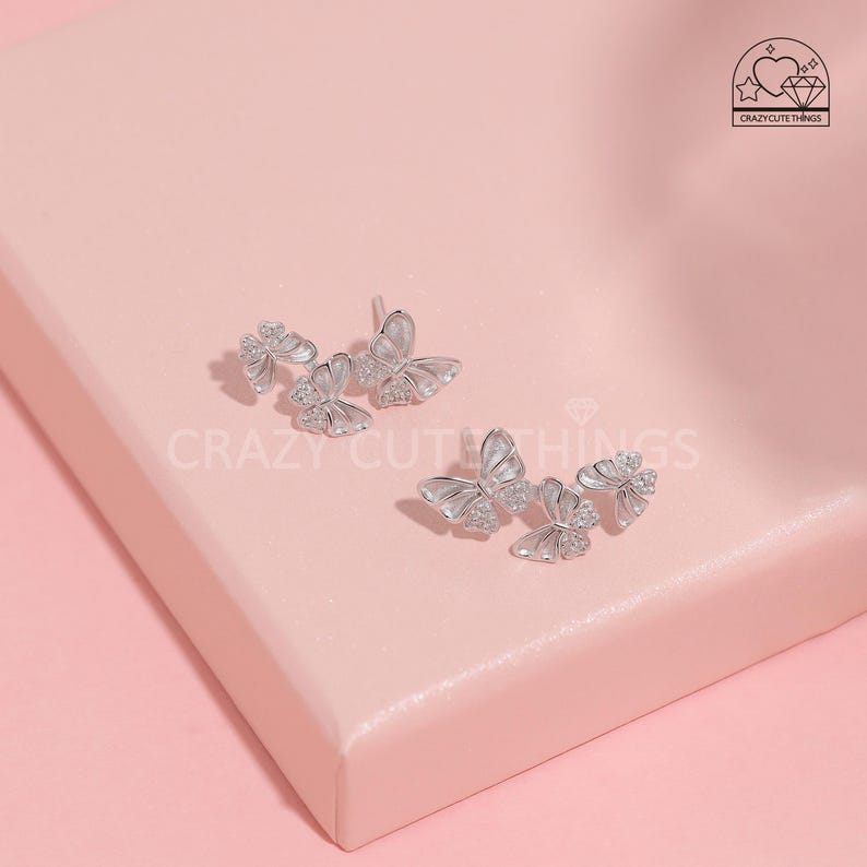 May include: A pair of silver butterfly earrings with clear crystal accents. Each earring features three butterflies, with the largest butterfly in the centre and two smaller butterflies on either side. The butterflies are arranged in a staggered pattern, creating a delicate and feminine look.