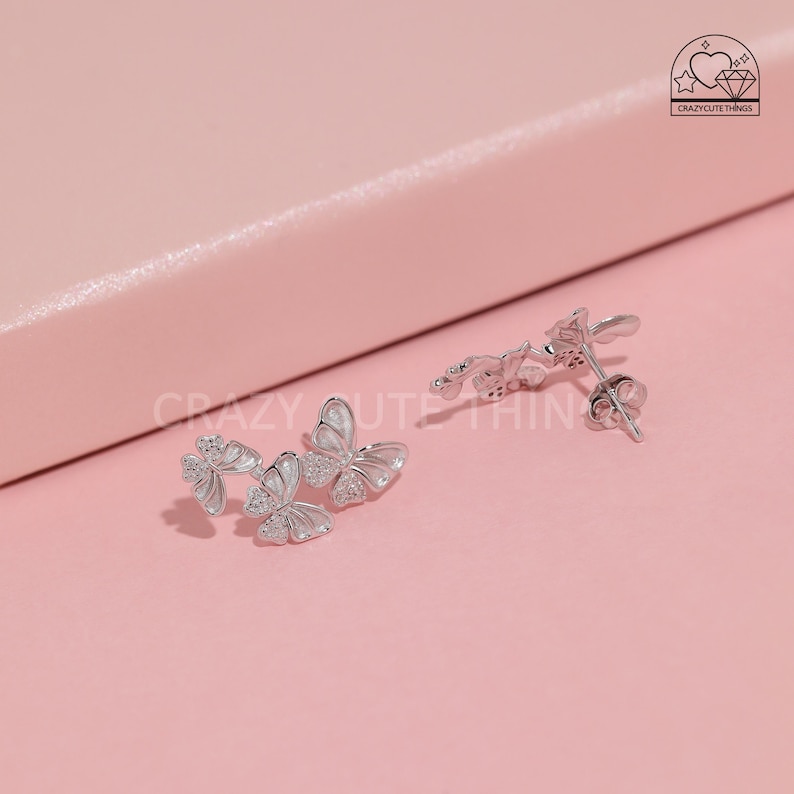 May include: A pair of silver butterfly earrings with clear crystals. The earrings are on a pink surface.