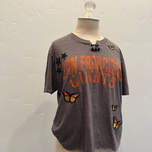 May include: A gray t-shirt with the text "San Francisco Giants" in orange. The shirt has a butterfly and star embellishments.