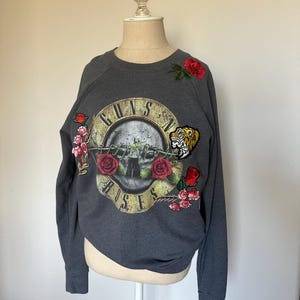 May include: Dark gray sweatshirt featuring the Guns N' Roses logo with a skull and crossbones design. The sweatshirt has embroidered red roses and a tiger patch. The long-sleeved top is displayed on a mannequin.