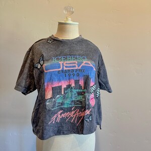 May include: A cropped, gray, t-shirt with a vintage-style graphic print of a race car and the text "ICEBERG USA GRAND PRIX 1990" and "Phoenix Arizona". The shirt has several butterfly and other small graphic embellishments.