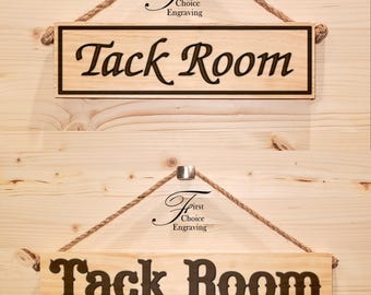 Può includere: Un cartello in legno con il testo "Tack Room" in un carattere decorativo. Il cartello è appeso a una corda con un gancio di metallo. Il cartello è in legno chiaro e ha una macchia marrone scuro.