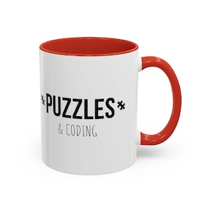 Può includere: Tazza in ceramica bianca con bordo e manico rossi. La tazza ha un testo nero che recita "*PUZZLES* & CODING"