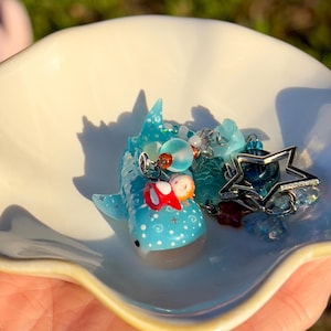 May include: A light blue whale shark figurine with white polka dots and star accents, featuring a small orange-haired figure in a red outfit on its back. The shark is attached to a silver-toned star charm and various blue and clear beads.