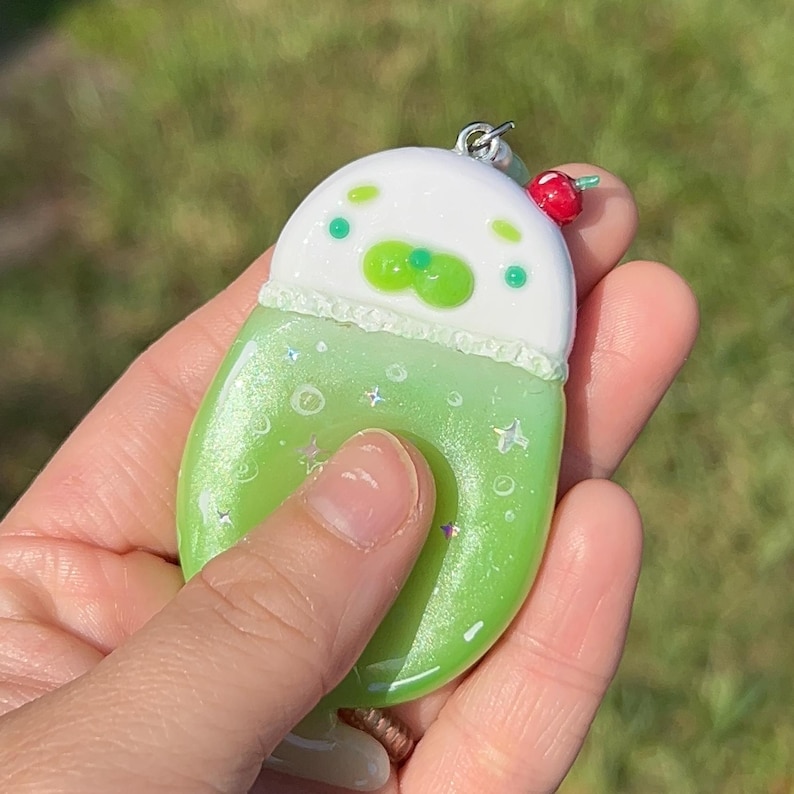 May include: A resin charm shaped like a drink with a white top and a light green base. The white section features green dots and a small red cherry accent. The green base has a glittery finish with small white star and circle details.