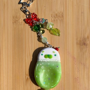 May include: A keychain featuring a star-shaped metal clasp attached to a charm shaped like a green drink in a glass with a white top, decorated with small beads, a red butterfly, and a green leaf charm hanging from silver-toned metal links.