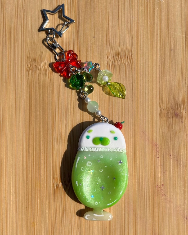 May include: A keychain featuring a star-shaped metal clasp attached to a charm shaped like a green drink in a glass with a white top, decorated with small beads, a red butterfly, and a green leaf charm hanging from silver-toned metal links.
