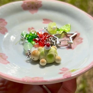 May include: A keychain featuring a light green turtle charm with a small white and red topper, accented by translucent green beads, a red flower-shaped charm, a bright green leaf charm, and a silver-toned star-shaped metal clasp attachment.