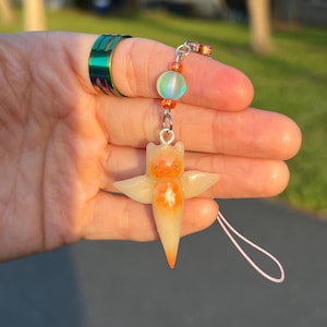 May include: A phone charm featuring a translucent, pale orange creature with small wings and a pointed base. The charm is attached to a silver-toned metal loop with a light blue iridescent bead and two small orange beads, suspended from a thin pink cord.