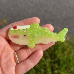 May include: A lime green whale-shaped resin charm featuring a white cream-like topping on its head with a small red cherry accent. The body is decorated with white circular bubble patterns and small holographic star-shaped glitter accents.