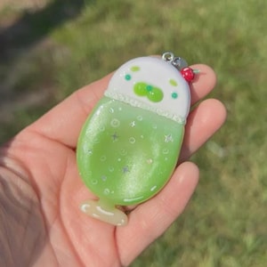 May include: A glossy, pearlescent green and white charm shaped like a drink in a glass. The top section is white with green dots, while the bottom is a light green with small star and circle accents. A small red bead is attached to the silver metal loop.