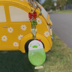 May include: A keychain featuring a red bow, green beads, and a white and green drink charm with a small red cherry on top. The charm is shaped like a glass with a green liquid, white foam, and star accents, hanging from a yellow car-shaped bag.