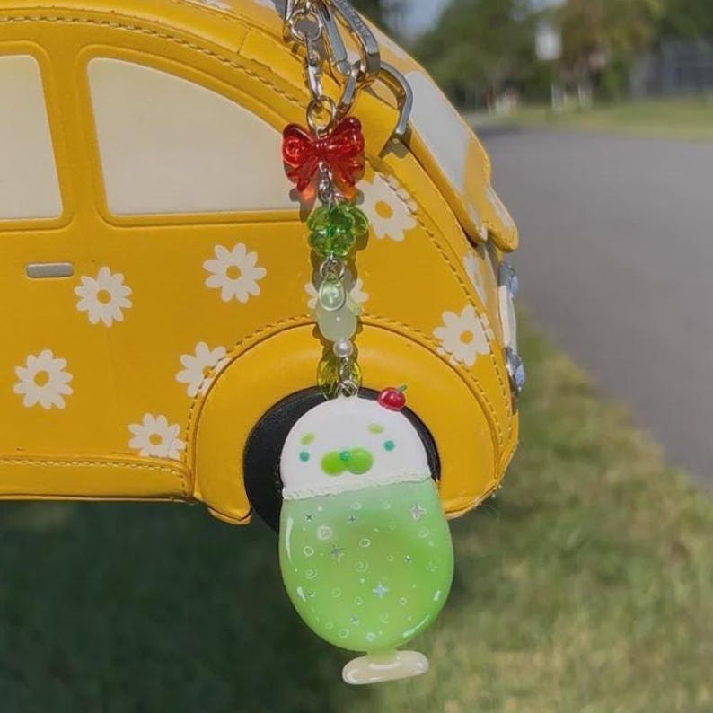 May include: A keychain featuring a red bow, green beads, and a white and green drink charm with a small red cherry on top. The charm is shaped like a glass with a green liquid, white foam, and star accents, hanging from a yellow car-shaped bag.