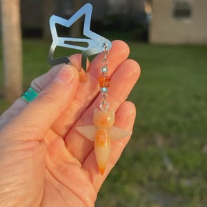 May include: A silver-toned star-shaped hair clip featuring a dangling charm. The charm consists of small light blue and orange beads connected to a translucent, pale orange, winged creature figurine with a pointed base and subtle star detail.