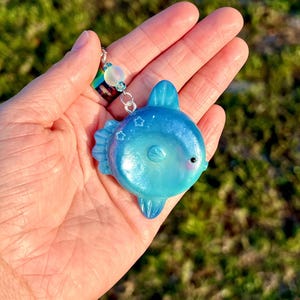 May include: A glossy, iridescent blue sunfish charm featuring a smooth, rounded body, a small black eye, and subtle star-shaped engravings. The charm is attached to a silver-toned metal loop with a single translucent, opalescent bead.
