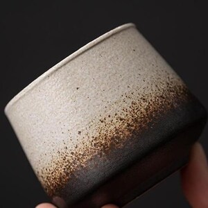 Könnte beinhalten: Eine Keramik-Tasse mit strukturierter Oberfläche. Die Tasse verläuft von hellem Beige oben zu dunklem Braun am Boden, mit gesprenkelten Details. Die Tasse wird von einer Hand vor dunklem Hintergrund gehalten.