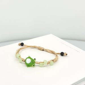 May include: A bracelet featuring a central green glass frog charm with a white belly, flanked by light green translucent beads on a tan woven cord. The adjustable cord ends are finished with small black beads, creating a simple, functional design.