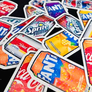 May include: A collection of holographic stickers featuring various soda can designs, including Coca-Cola, Fanta, Sprite Remix, and Pepsi. Each sticker has a die-cut white border and a shimmering, multi-colored glitter finish that reflects light.