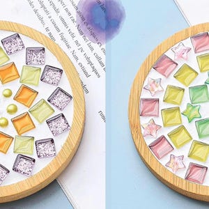May include: Two decorative coasters with a mosaic design. One features orange, yellow, and purple square tiles, a star, and pearl accents. The other has pink, yellow, and green tiles and star shapes, all set in a wooden frame.