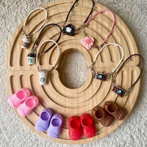 May include: Miniature accessories including coffee cup charms, flower and camera pendants, and pairs of shoes in pink, purple, red, and brown. The items are arranged on a wooden, circular tray.