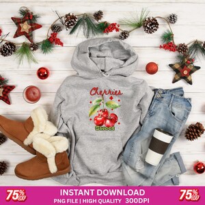 May include: A gray hoodie with a cherry graphic and the word "Cherries" in red. The image includes blue jeans, brown boots, and holiday decorations like pinecones and ornaments.