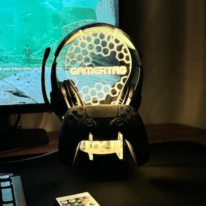 May include: A black gaming headset and controller stand with a clear acrylic base and a honeycomb design. The stand has a yellow light that illuminates the word "GAMERTAG" on the headset. The stand is on a black desk with a keyboard and a remote control.