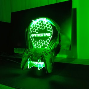 May include: A green gamertag light with a honeycomb design and the word "GAMERTAG" in the center. The light is mounted on a stand with a camouflage-patterned video game controller.