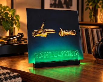 Op de afbeelding: Een rechthoekige acryl displaystandaard met een donkerblauwe vinyl platenhoes met een gouden handgebaar-afbeelding. De zwarte voet verlicht het heldere acrylpaneel met een felgroen licht en toont de tekst NOW PLAYING.