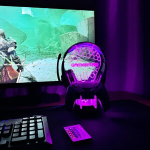 May include: A purple and white gaming headset lamp with the word "GAMERTAG" printed on the front. The lamp is on a black desk with a keyboard and a remote control.