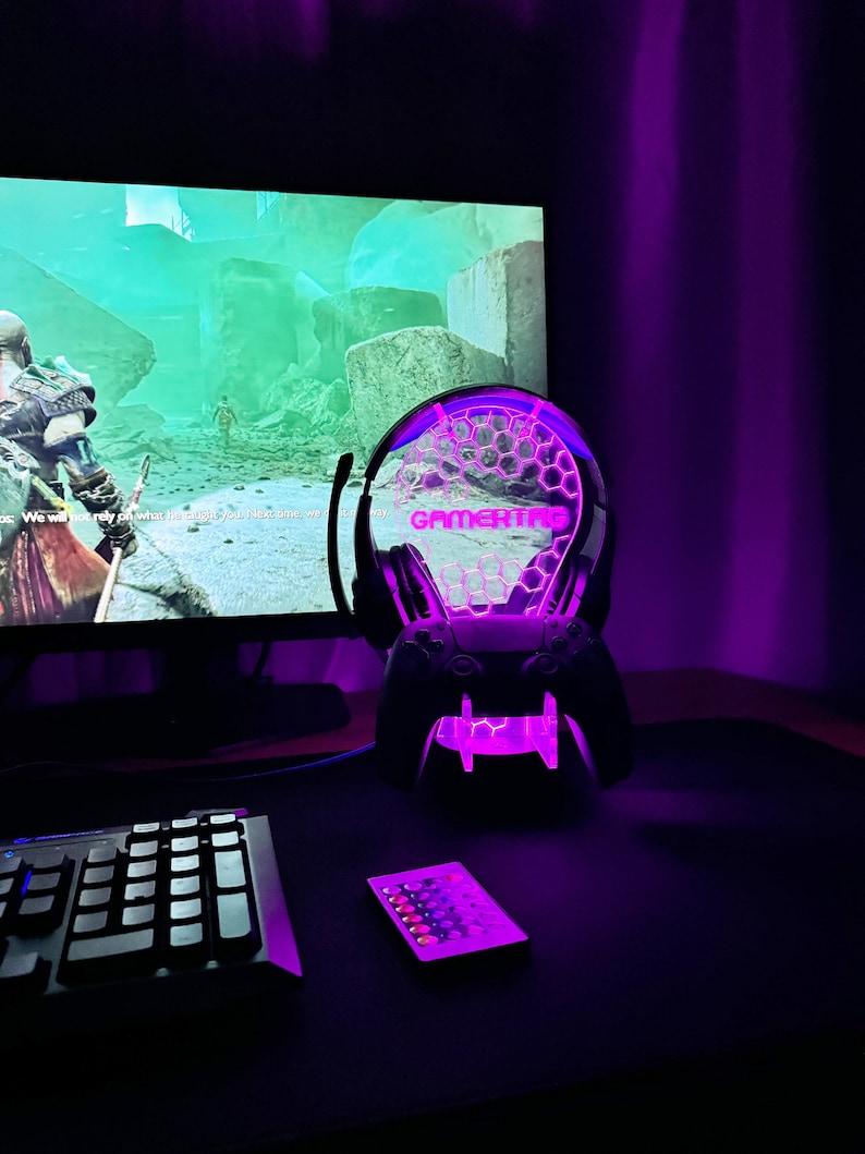 May include: A purple and white gaming headset lamp with the word "GAMERTAG" printed on the front. The lamp is on a black desk with a keyboard and a remote control.