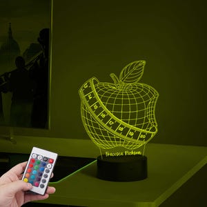May include: A yellow 3D printed night light shaped like an apple with a measuring tape around it. The apple is missing a bite and the measuring tape reads from 13 to 20. The base of the night light is black and has the name "Francesca Richardson" printed on it.