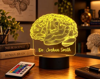 Puede incluir: Una lámpara LED amarilla con un diseño de cerebro humano grabado en un panel acrílico transparente. La base es negra y cilíndrica. El texto "Dr. Joshua Smith" está grabado debajo del cerebro. Incluye un pequeño mando a distancia.